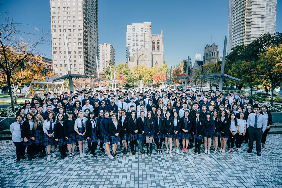 Un gruppo di studenti della Pattison High School si raduna davanti a un panorama urbano con edifici alti e un campanile.