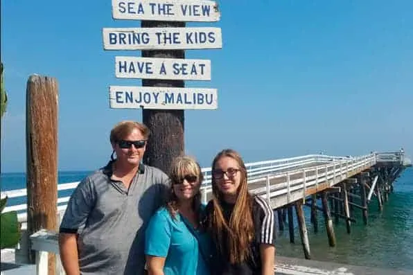 Tres personas posan sonrientes junto a un cartel de madera con un muelle al fondo