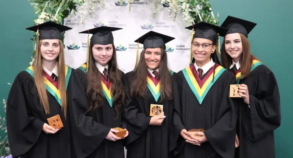 Cinque studenti della Queen Margaret’s School indossano la toga di laurea e tengono i loro diplomi davanti a un logo universitario.