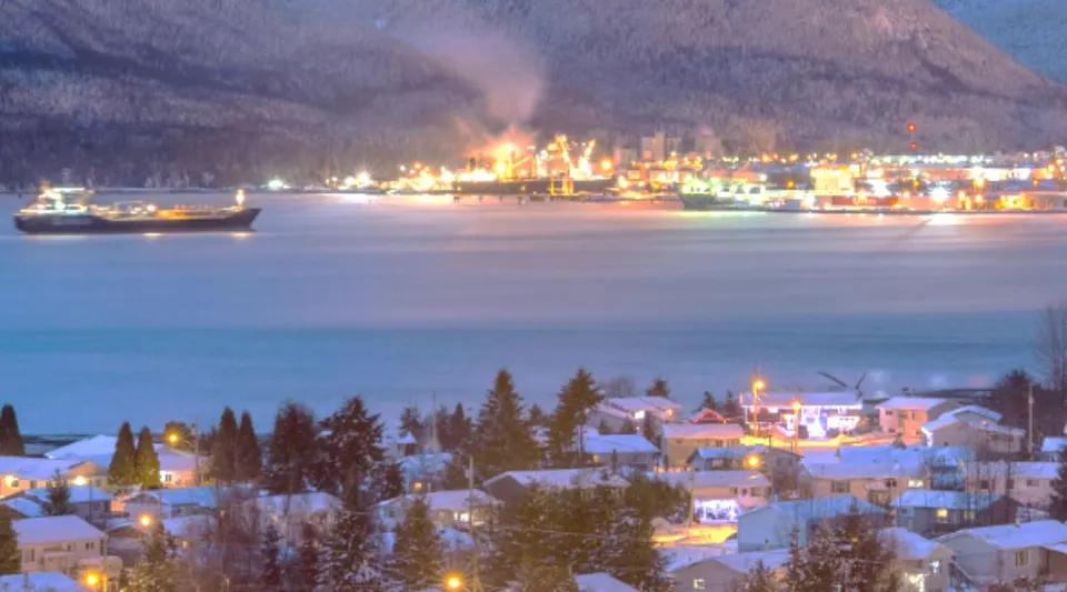 Case coperte di neve a Kitimat con vista sull'acqua, una nave e impianti portuali illuminati davanti alle montagne.
