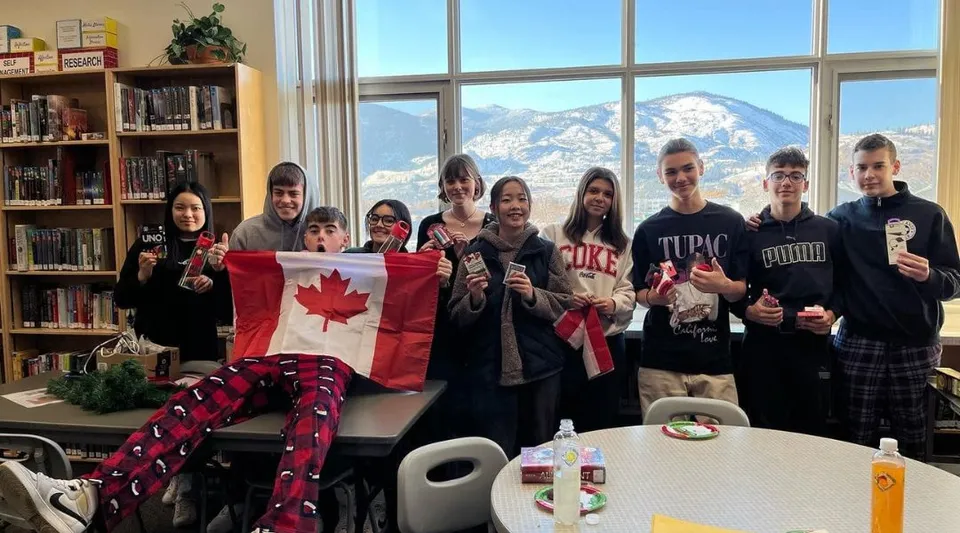 Un gruppo di studenti della Princess Margaret Secondary School tiene una bandiera canadese in una sala comune con vista su montagne innevate.