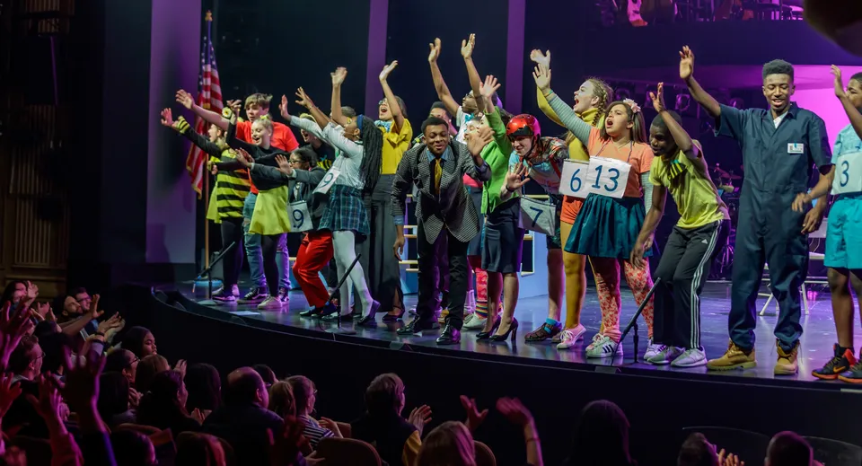 Studenti della Bournemouth Collegiate School mostrano una vivace performance sul palco con le braccia alzate in una scenografia multicolore.