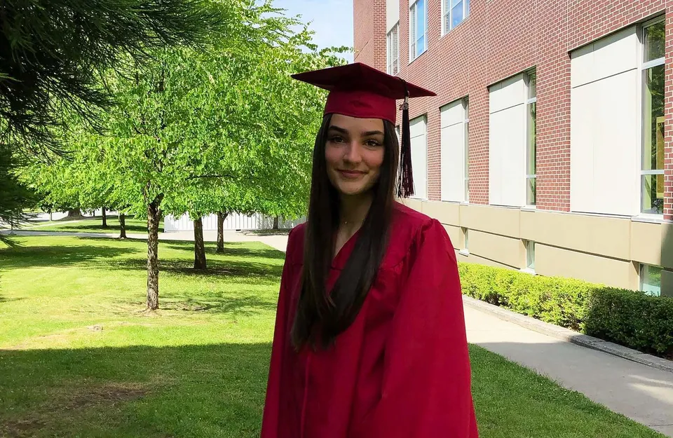 Una giovane donna in toga da diploma rossa sta in piedi davanti all'edificio in mattoni della Lord Byng Secondary School.
