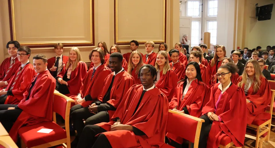 Studenti della St. Edmunds School siedono durante una solenne cerimonia di diploma in una stanza con grandi finestre e decorazioni magnifiche.