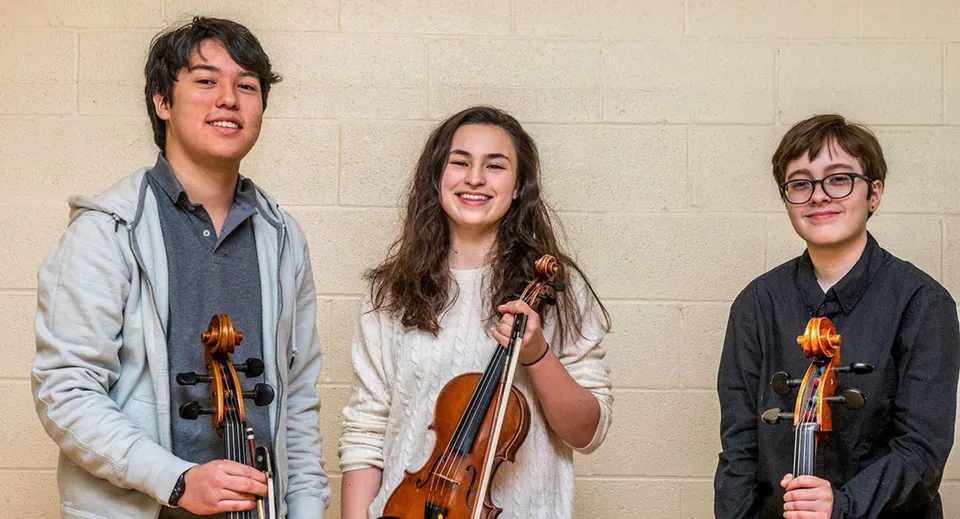 Tre studenti della Eagle School tengono in mano strumenti musicali e si trovano davanti a una parete semplice.