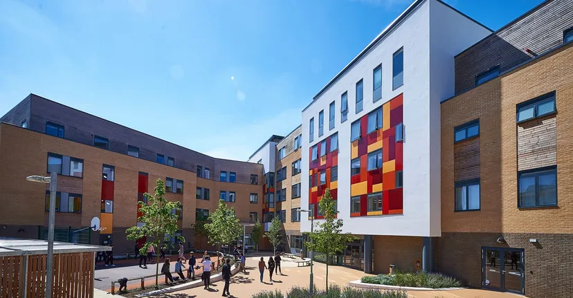 L'edificio moderno a più piani con motivi geometrici colorati mostra l'Abbey College Cambridge, in primo piano si vedono alberi e persone.