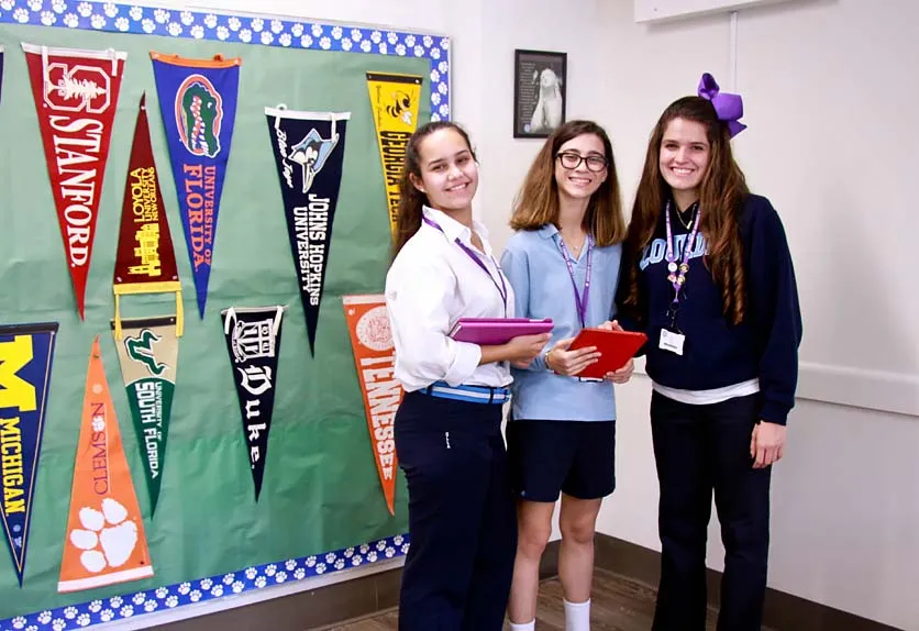 Tres estudiantes están de pie en una habitación con banderines escolares de colores y pancartas en las paredes de la Lourdes Academy.