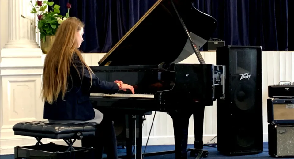 Una donna con lunghi capelli biondi suona il pianoforte sul palco del Midleton College davanti a un sipario nero.