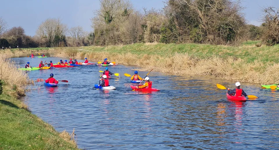 Studenti del Beech Hill College pagaiano su kayak colorati su un fiume con una verde vegetazione sulle sponde.