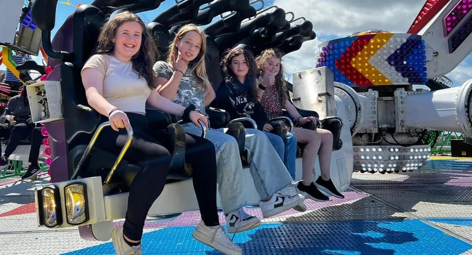Studentesse della Monmouth School for Girls siedono insieme su una giostra in un parco divertimenti con attrazioni colorate sullo sfondo.