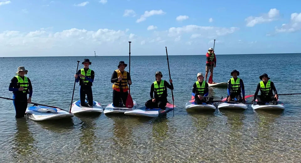 Studenti del Plympton International College stanno su stand-up paddle con giubbotti di salvataggio nell'acqua bassa davanti a un oceano calmo.