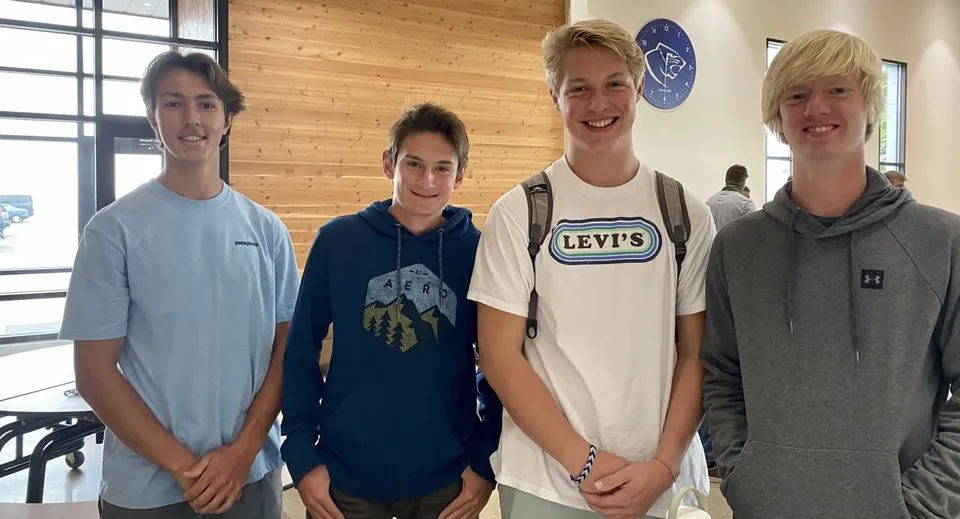 Quattro studenti posano insieme davanti a una parete con logo nel campus della Stillwater Christian School.