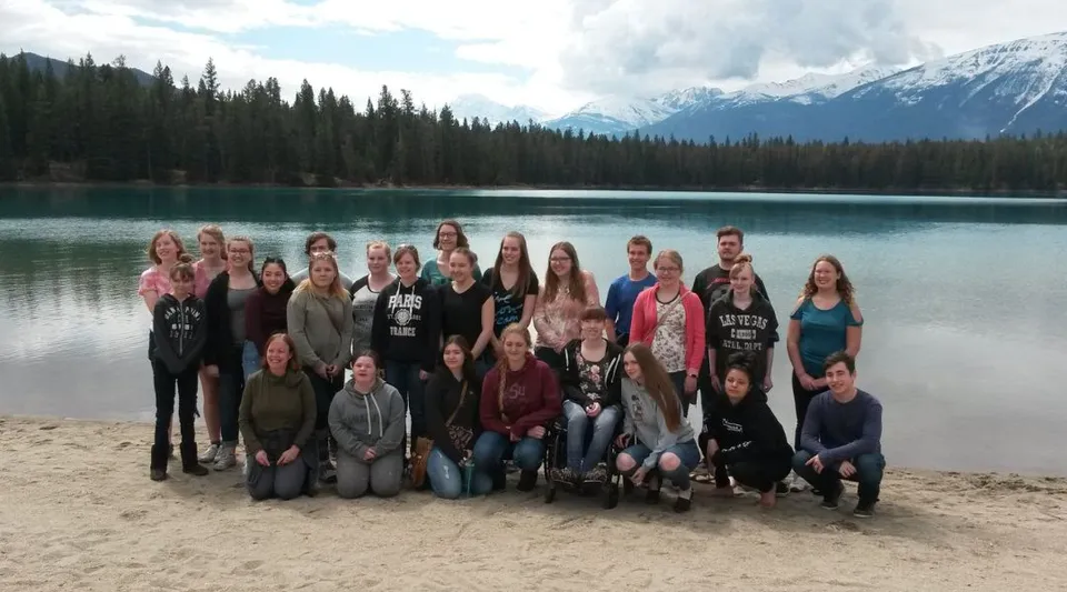 Studenti della New Sarepta Community High School si radunano su una spiaggia sabbiosa davanti a un lago con montagne innevate sullo sfondo.