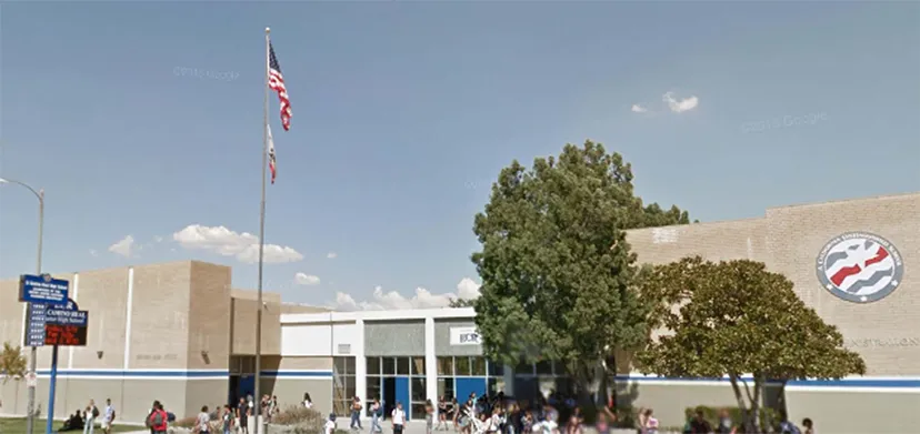 La bandiera americana sventola sopra l'edificio a più piani della El Camino Real Charter High School, con alberi e persone in primo piano.