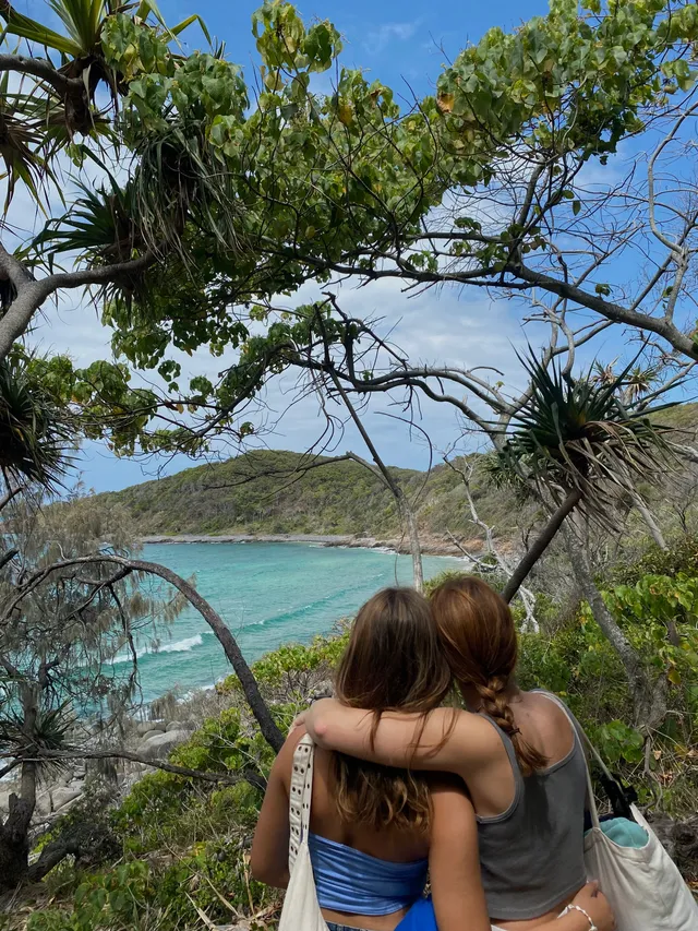 Dos chicas abrazadas de espaldas contemplando una cala de aguas turquesas entre la vegetación