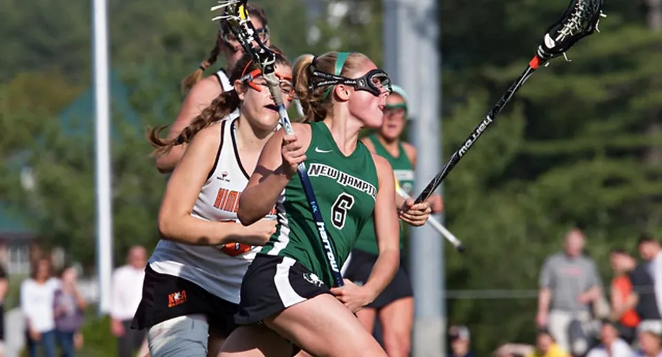 Due giocatrici di lacrosse della New Hampton School si sfidano su un campo sportivo davanti al pubblico.