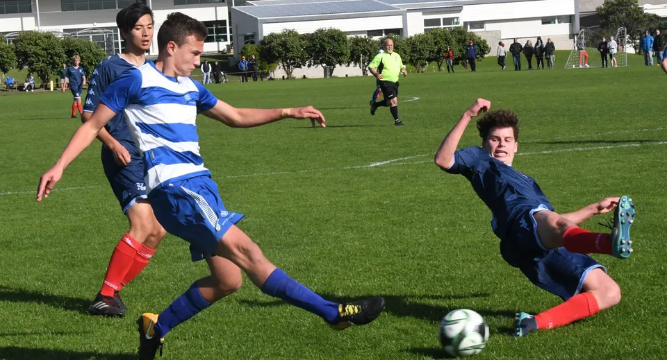 Due calciatori della Onehunga High School si contendono la palla sul campo di gioco, mentre altri giocatori e un arbitro sono visibili sullo sfondo.