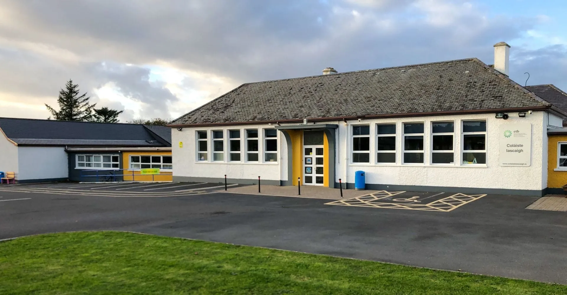 L'edificio scolastico a un piano dell'Iascaigh College con porta d'ingresso gialla, circondato da un'area pavimentata e un prato.