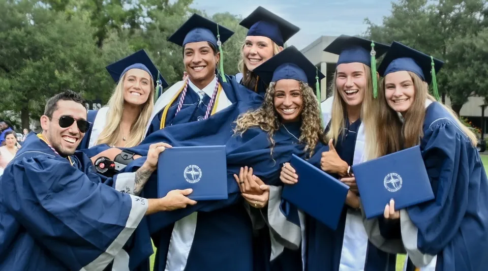 Un gruppo di laureate e laureati della University of North Florida in piedi all'aperto con toghe e tocchi blu, circondati da alberi.