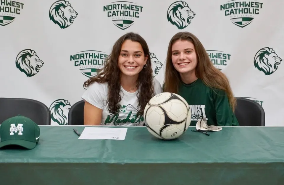 Due studentesse della Northwest Catholic High School siedono a un tavolo con un pallone da rugby davanti a uno sfondo con logo.