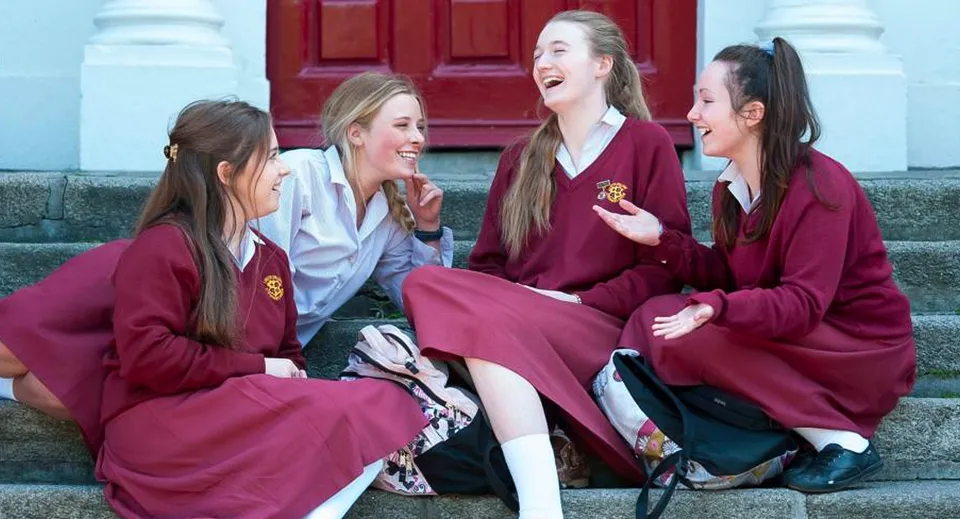 Le studentesse del Loreto College, St. Stephen's Green, sorridono in toga rossa davanti a una porta di legno rossa.