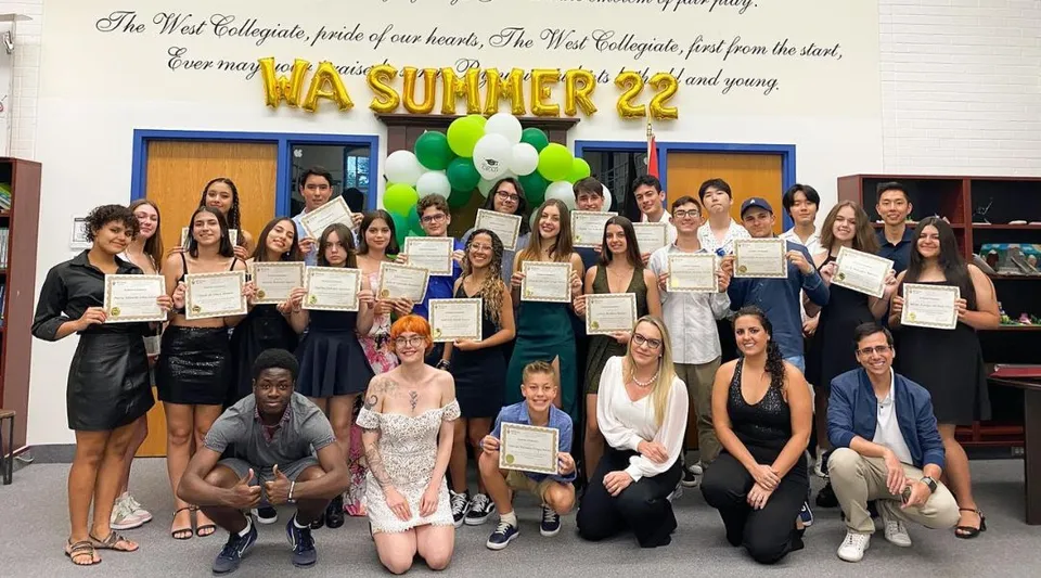 Un gruppo di studenti e studentesse della William Academy è in piedi con dei certificati davanti a un banner della festa estiva 2022.