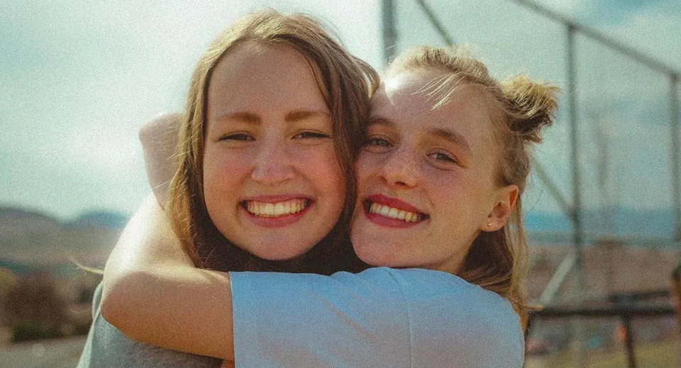 Due studentesse con i capelli biondi si abbracciano sorridendo davanti a un paesaggio naturale nel campus della Vernon Secondary School.