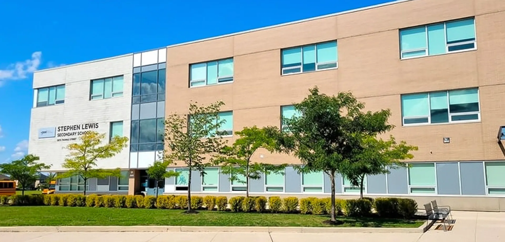 L'edificio a più piani della Stephen Lewis Secondary School con grandi finestre si trova davanti a un cielo azzurro limpido e ad alberi.