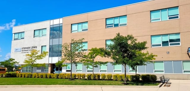 Das mehrstöckige Gebäude der Stephen Lewis Secondary School mit großen Fenstern steht vor klarem blauem Himmel und Bäumen.