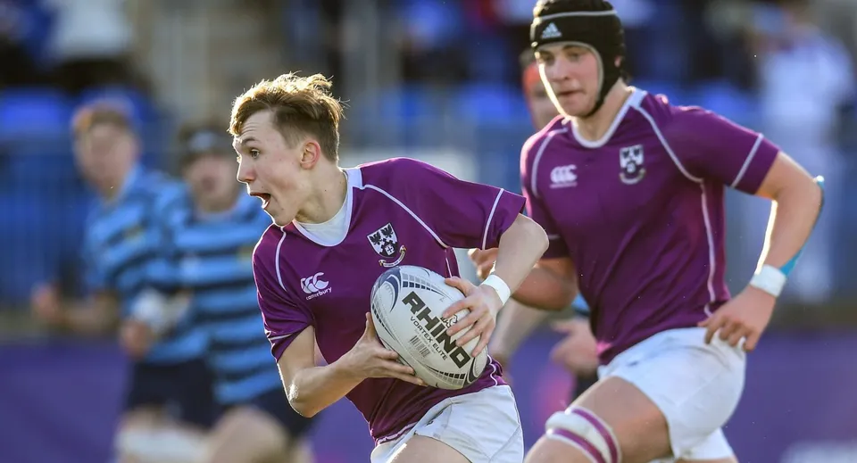 Due giocatori di rugby del Clongowes Wood College in maglia viola corrono sul campo da gioco con la palla.