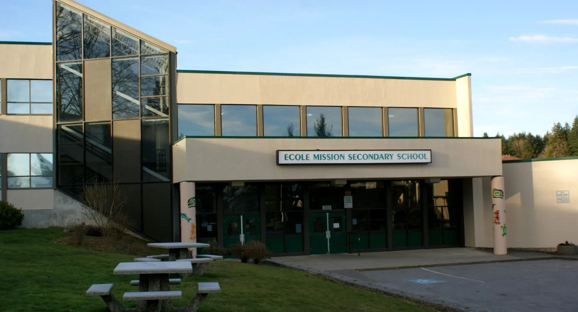 L'edificio scolastico moderno della Mission Secondary School con facciata in vetro e un'area verde con panchine in primo piano.