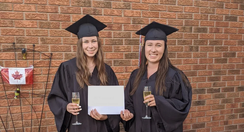 Due studentesse della Longfields-Davidson Heights Secondary School posano con diplomi e calici di spumante davanti a un muro di mattoni con la bandiera canadese.