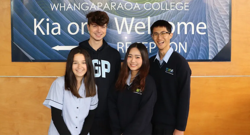 Un gruppo di quattro studenti posa davanti a uno striscione con la scritta 'Kia ora Welcome' nel campus del Whangaparāoa College.