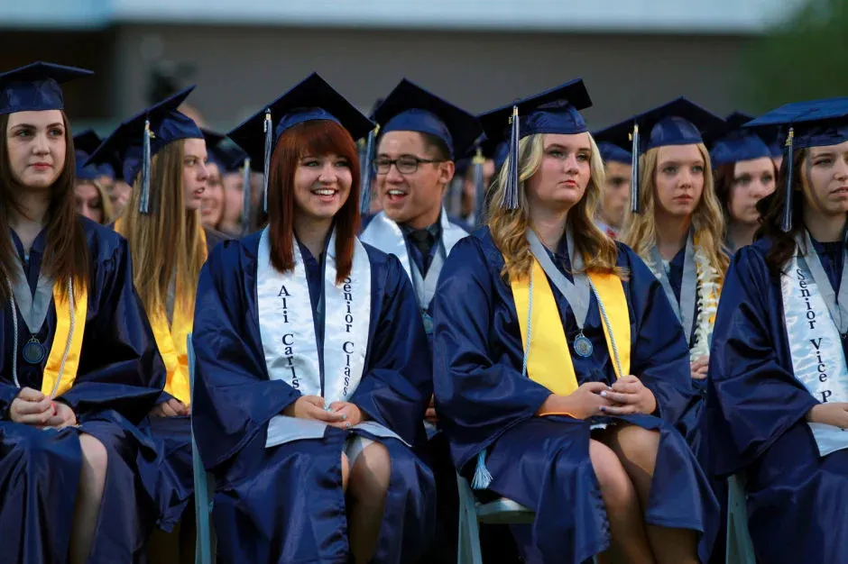 Estudiantes del Higley Unified School District llevan túnicas de graduación azules y amarillas y están sentados juntos en un auditorio.