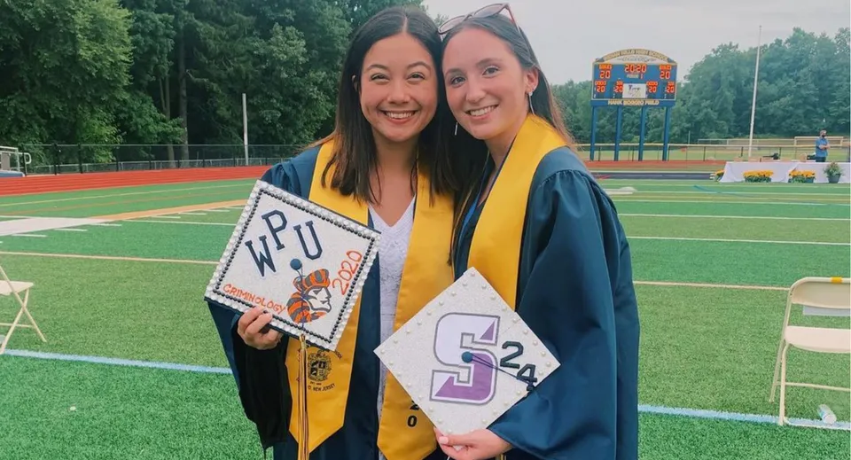 Due studentesse del Las Virgenes Unified School District in abiti da diploma sono in piedi su un campo sportivo mentre tengono cartelli di laurea.