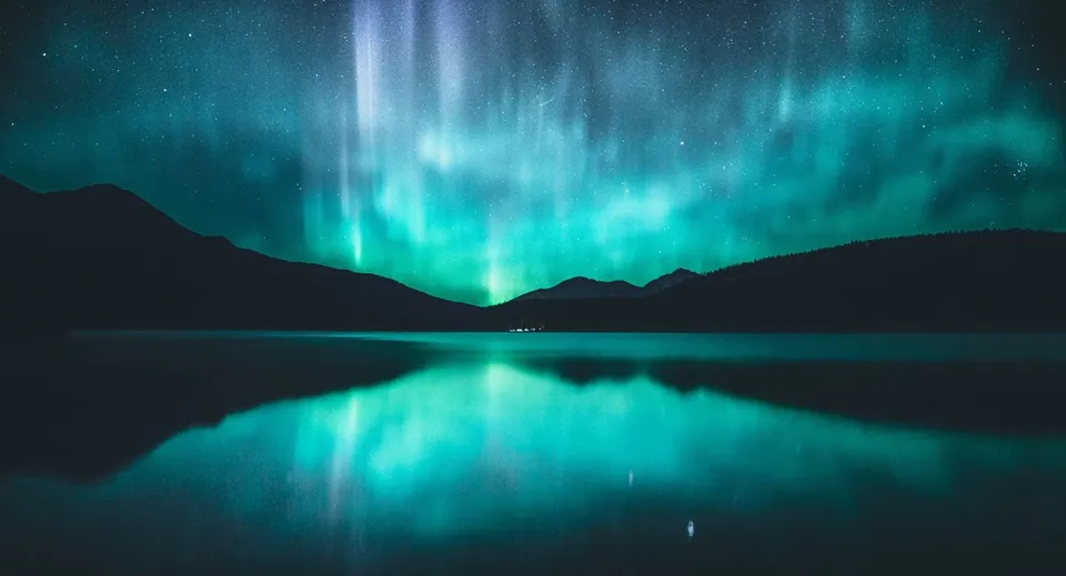 Un'aurora boreale verde brilla sopra un lago calmo e si riflette nell'acqua davanti a sagome di montagne.