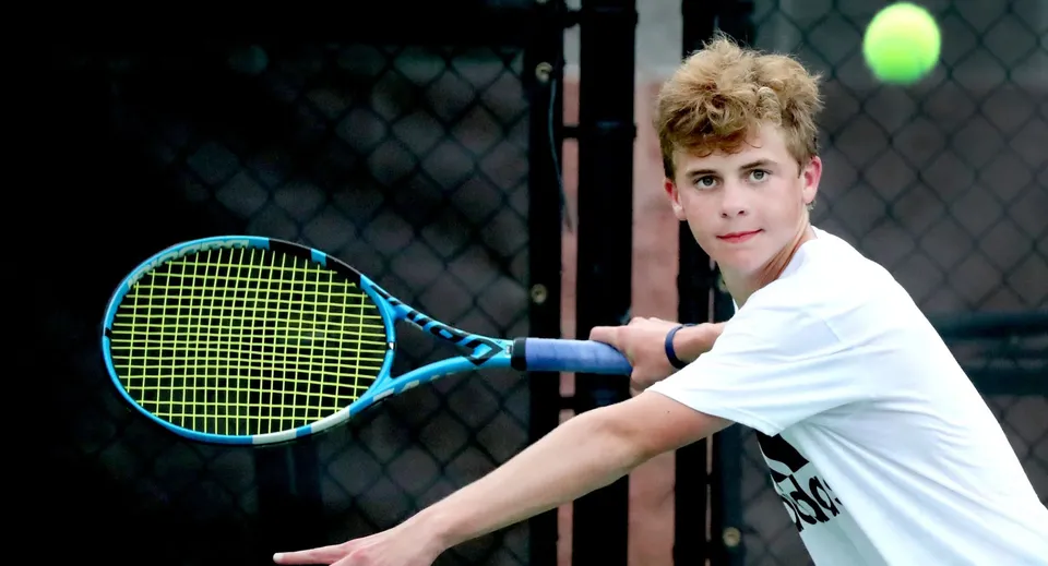 Uno studente della Merrick Preparatory School si trova sul campo da tennis con una racchetta davanti alla recinzione metallica.