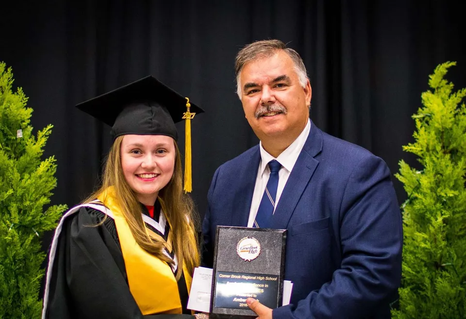 Una diplomata si trova accanto a un uomo in abito elegante davanti a uno sfondo verde nel campus della Corner Brook Regional High School.