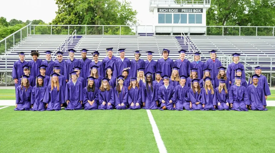 Un gruppo di studenti della Norfolk Christian Schools indossa le toghe di diploma viola su un campo davanti a una struttura simile a uno stadio.