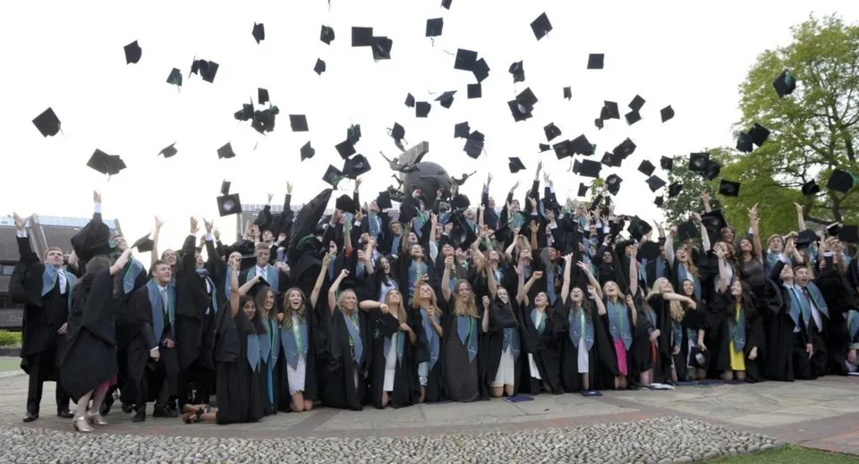 Un gruppo di diplomati lancia i propri cappelli in aria nel campus del Bradfield College davanti ad alberi verdi ed edifici.