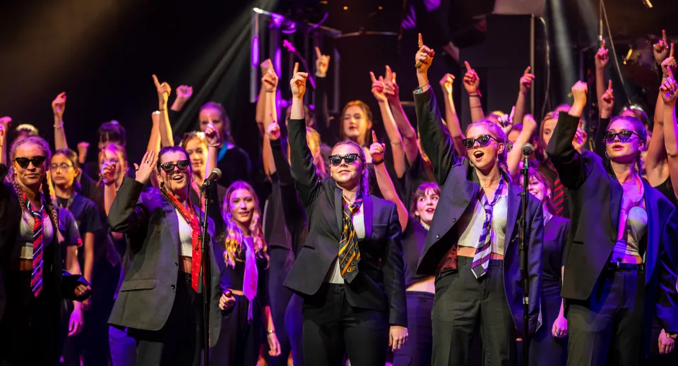 Un gruppo di studenti della West Buckland School si trova sul palco a braccia alzate sotto luci colorate.
