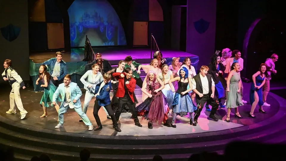 Un gruppo di studenti e studentesse in costumi colorati si trova sul palco della M.M. Robinson High School davanti a uno sfondo astratto.