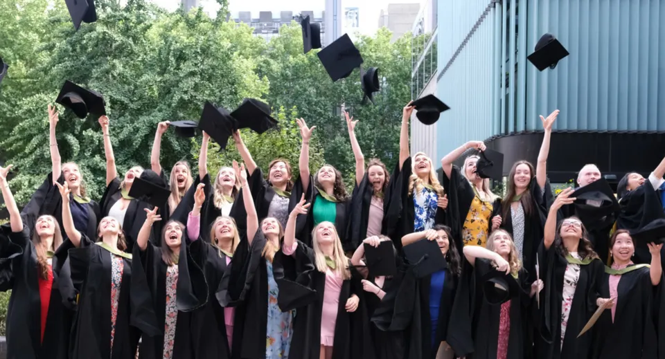 Un gruppo di diplomati della Bloxham School lancia i propri tocchi in aria durante la cerimonia di diploma davanti ad alberi ed edifici.
