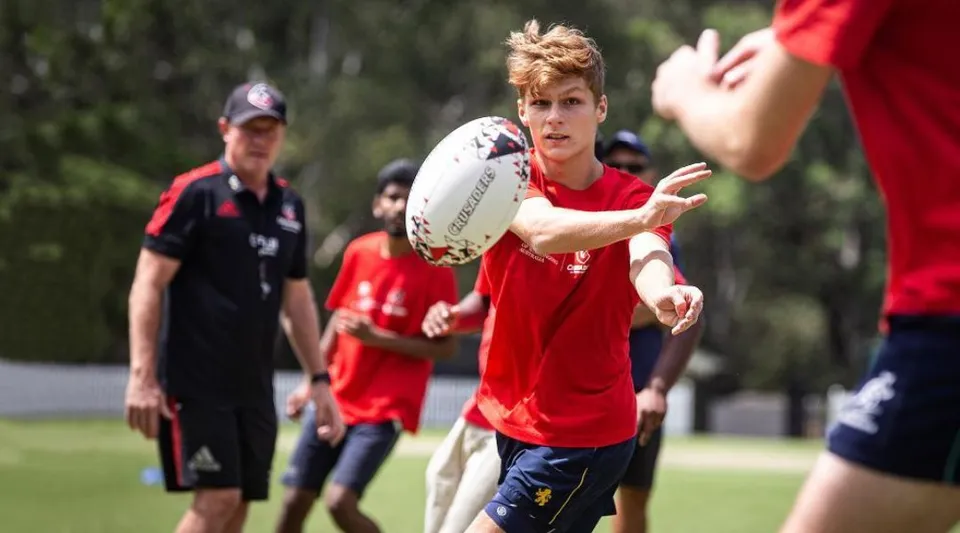 Un giovane in maglia rossa corre con un pallone da rugby sul campo da gioco nel campus della Wollongong University.