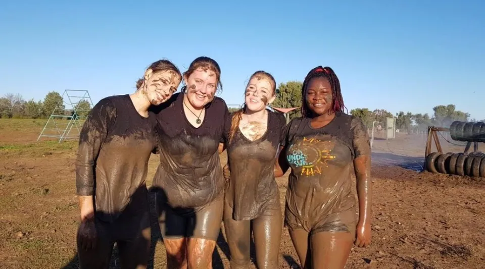 Un gruppo di quattro studentesse si trova in un campo sotto un cielo blu e alberi nel campus della Toormina High School.