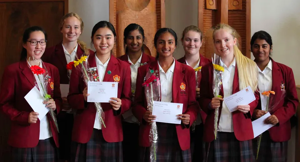 Le studentesse del Sacred Heart Girls' College Hamilton indossano stole di diploma rosse e tengono in mano i certificati in una stanza con pannelli in legno.