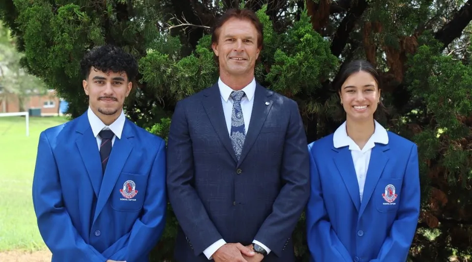 Tre persone, tra cui due studenti in divisa blu, si trovano davanti a uno sfondo verde all'interno del campus della Port Hacking High School.