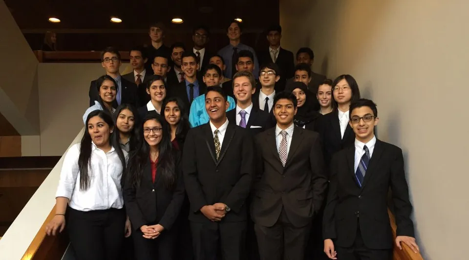 Un gruppo di studenti e studentesse della Garth Webb Secondary School sta in abito formale davanti a una scala di legno.