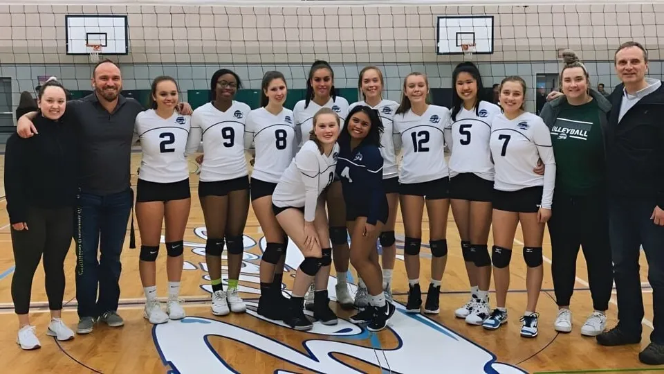Estudiantes de la Riverside Secondary School están de pie con sus uniformes de voleibol en una cancha de baloncesto con canastas al fondo.