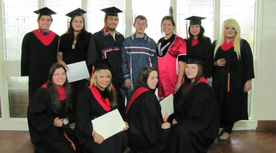 Un gruppo di diplomati e diplomate della Cornwall Collegiate & Vocational School in posa in abito accademico davanti a grandi vetrate.
